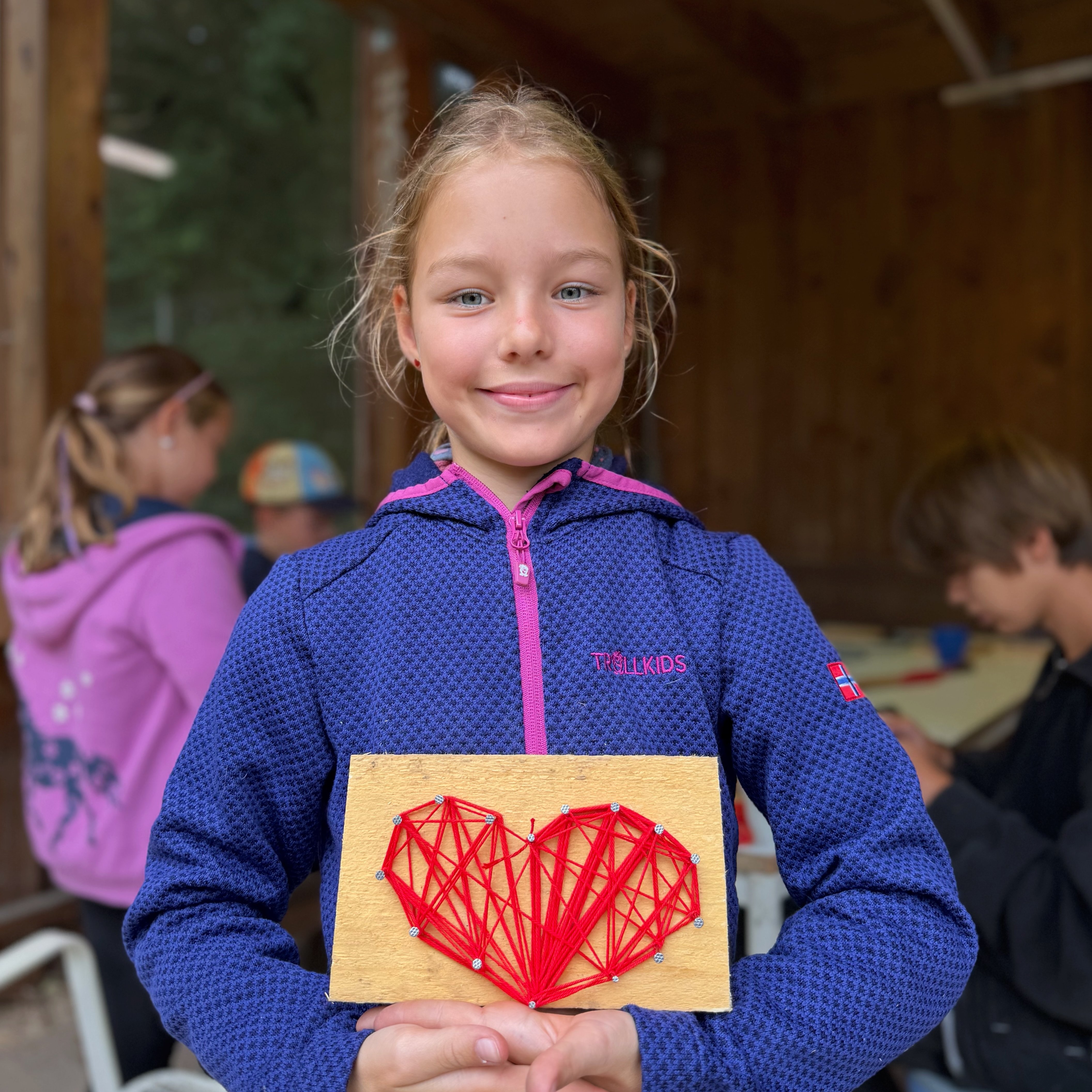 Ein Mädchen zeigt ein selbstgebasteltes rotes Herz. Es ist auf einem Holzbrett mit rotem Faden um Nägel gewickelt.